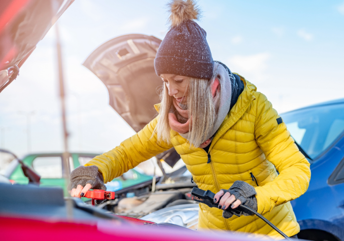 Woman in yellow coat and knit hat jump starting a car with another car in the background