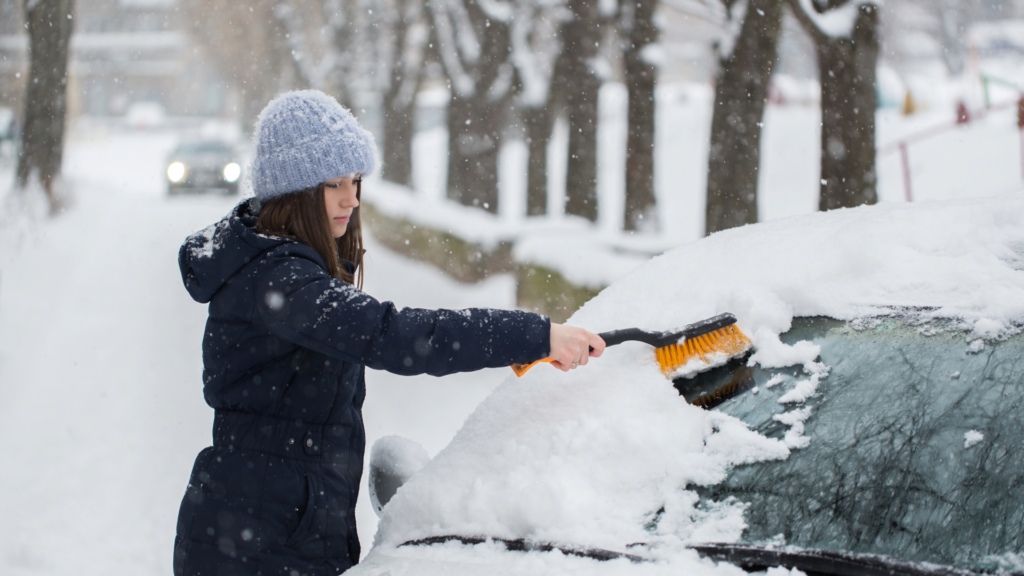 ounger teen driver in a hat and jacket using a snowbrush to clean off the windshield of her car.