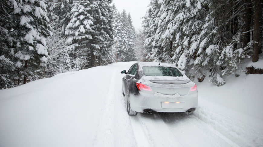 Snow-covered car traveling down a snow-covered road surrounded by pine trees.