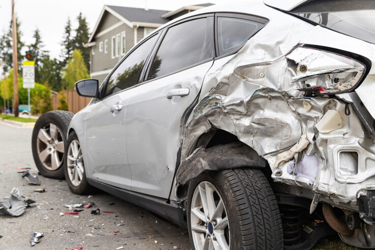 A silver car with damage to the rear fender, a broken axle and another vehicle's tire on the side of a street